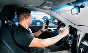 A distracted driver is using a cell phone while driving, one hand holding the phone and the other on the steering wheel, focusing on the screen instead of the road. 