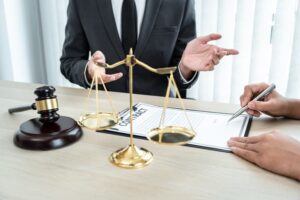 A male lawyer is consulting with a businesswoman client in his office. They are engaged in discussion, with legal documents and a laptop on the desk.