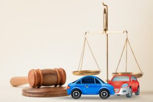 Two small toy car models, representing a crash, are placed on a desk in a courtroom alongside a judge's gavel. The setup symbolizes lawyer services, a civil court trial, or a vehicle accident case study.