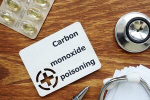 Wooden board with text reading 'Carbon monoxide poisoning' and a red cross silhouette, surrounded by a stethoscope, pills, blister packs, papers, and a wooden table.