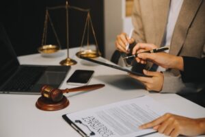 Image of a lawyer or legal advisor in a business suit, seated at a desk with an open legal book and contract documents in front of them. They are focused, holding a pen and appearing deep in thought, as if carefully reviewing and brainstorming the details of a business agreement. Nearby, a business partner looks on, symbolizing collaboration in ensuring accuracy in contract processing and discussing joint financial investment. The setting suggests a professional environment where attention to legal details and partnership are prioritized.