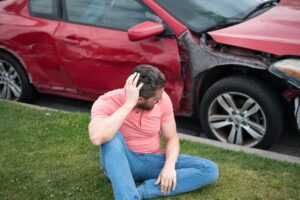 Young driver standing near a broken-down car on the road, clutching his head in shock and despair after an accident.