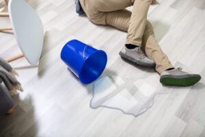 Mature man slipping on a wet floor at home, losing balance in front of a spilled bucket of water, with a surprised expression and arms flailing to break his fall.