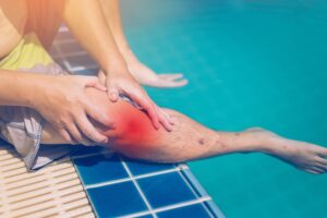 Swimmer sitting beside a swimming pool holding their injured knee, depicting an accident or injury near the pool area.