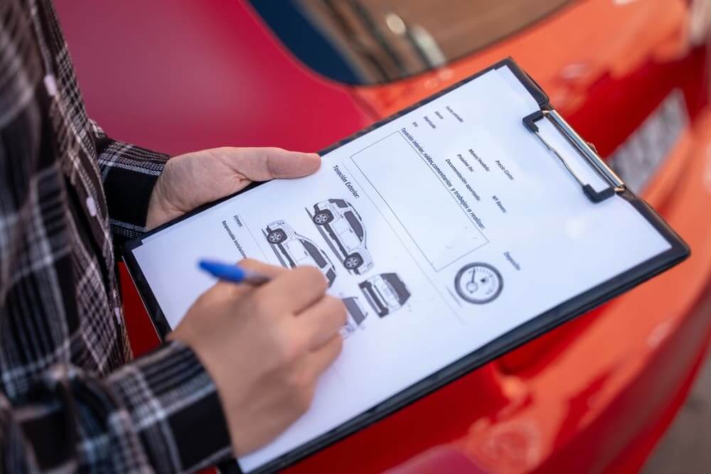 Insurance agent writing notes on clipboard, assessing damage on red car after traffic accident, filling insurance claim form