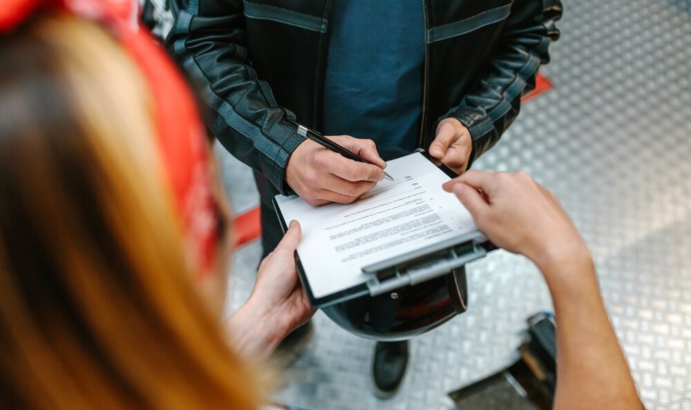 Biker signs insurance paperwork with female mechanic after accident, highlighting wage claims for time lost due to injuries.