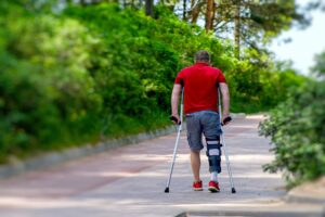 Injured man walking with crutches and leg brace on outdoor path during recovery.
